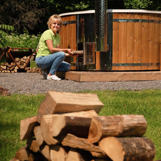 Frau in grünem Shirt und blauen Jeans legt Brennholz in den Ofen eines Hot Tubs im Garten
