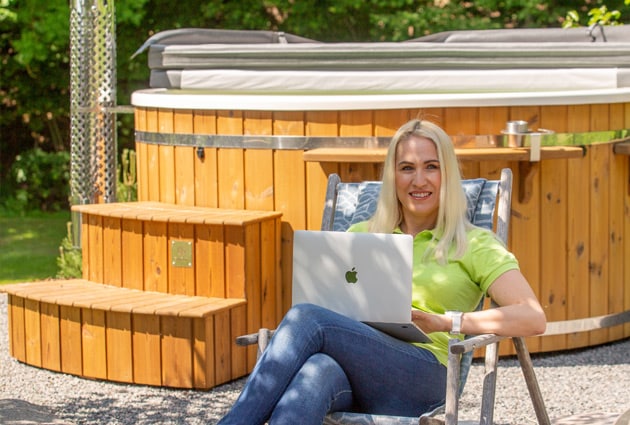 Woman sitting on outdoor chair using laptop in front of wood fired hot tub with wooden steps and cover closed