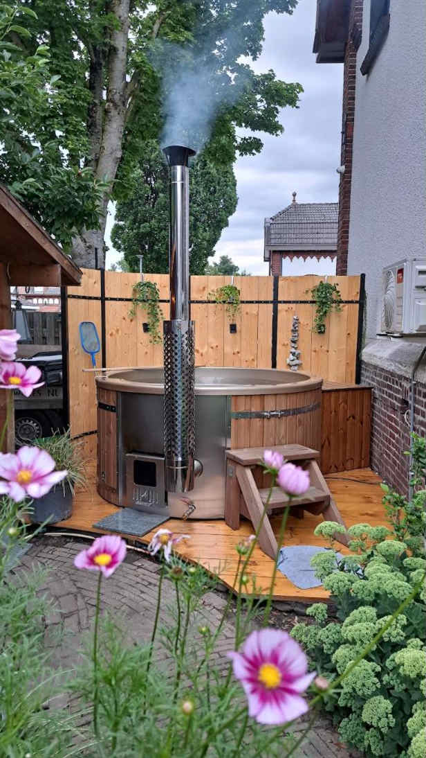 Außenansicht eines holzbefeuerten Whirlpools mit Metallkamin, Holztreppe und Blumengarten im Vordergrund
