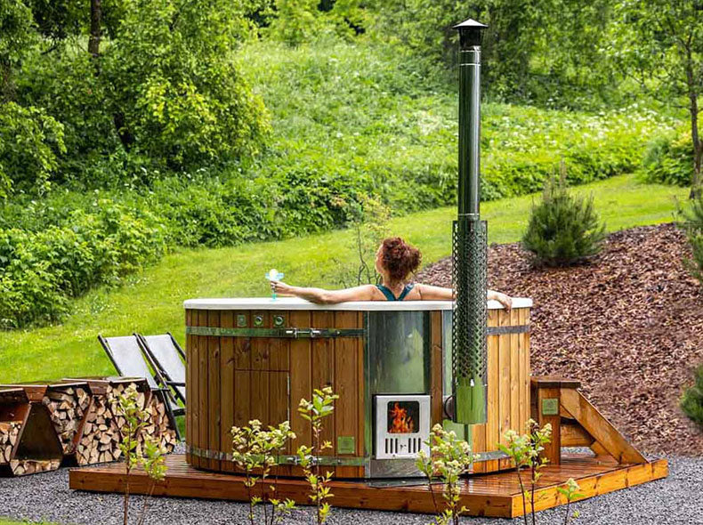Woman reclining alone in a wooden hot tub with wood-fired stove, holding a drink, set in a green garden.