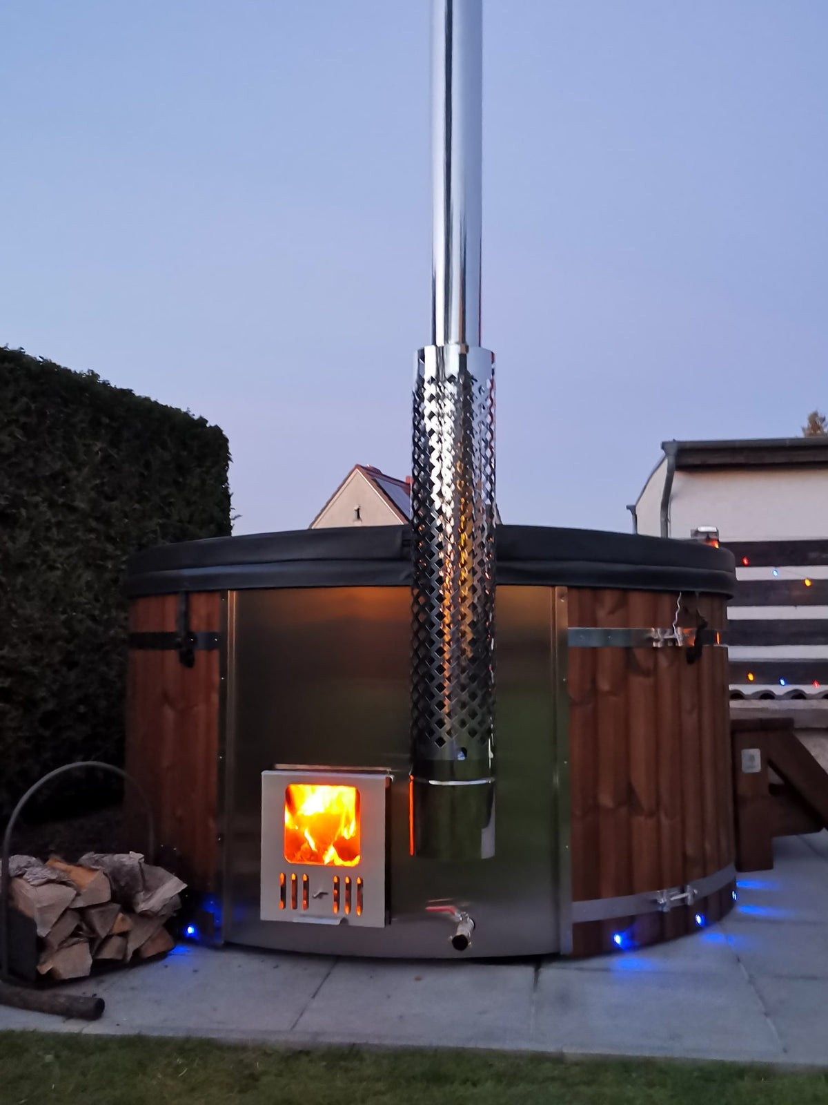 Außenansicht des holzbefeuerten hot tub mit Edelstahlofen, brennendem Feuer, hölzernen Außenverkleidungen und hohem Schornstein in der Abenddämmerung