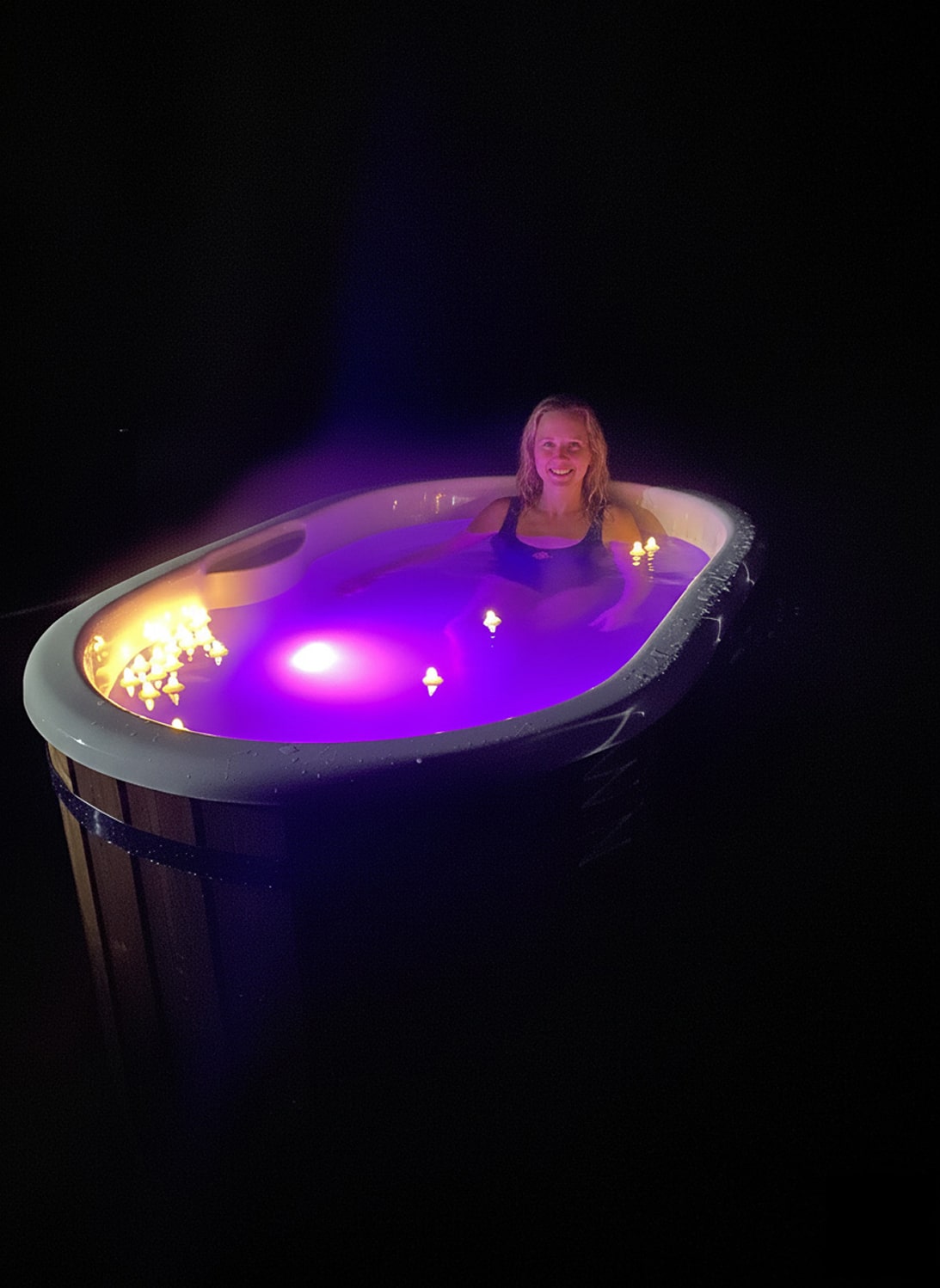 Nachtansicht einer Frau, die in einem beleuchteten ovalen Whirlpool mit violetten LED-Lichtern und schwimmenden Kerzen entspannt.
