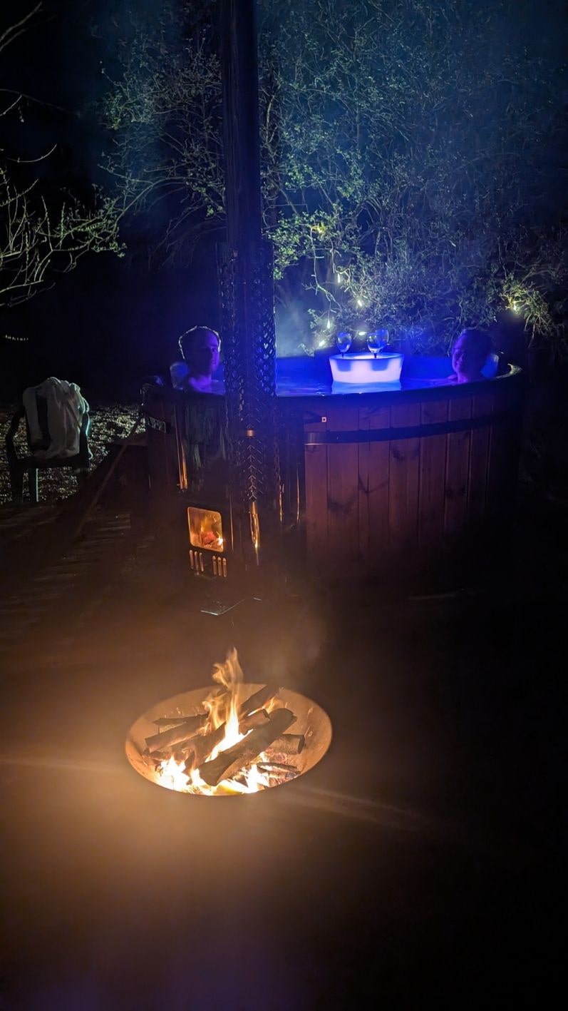 Nachtaufnahme von zwei Personen, die in einem holzbefeuerten Whirlpool neben einer leuchtenden Feuerstelle und Bäumen im Hintergrund entspannen.