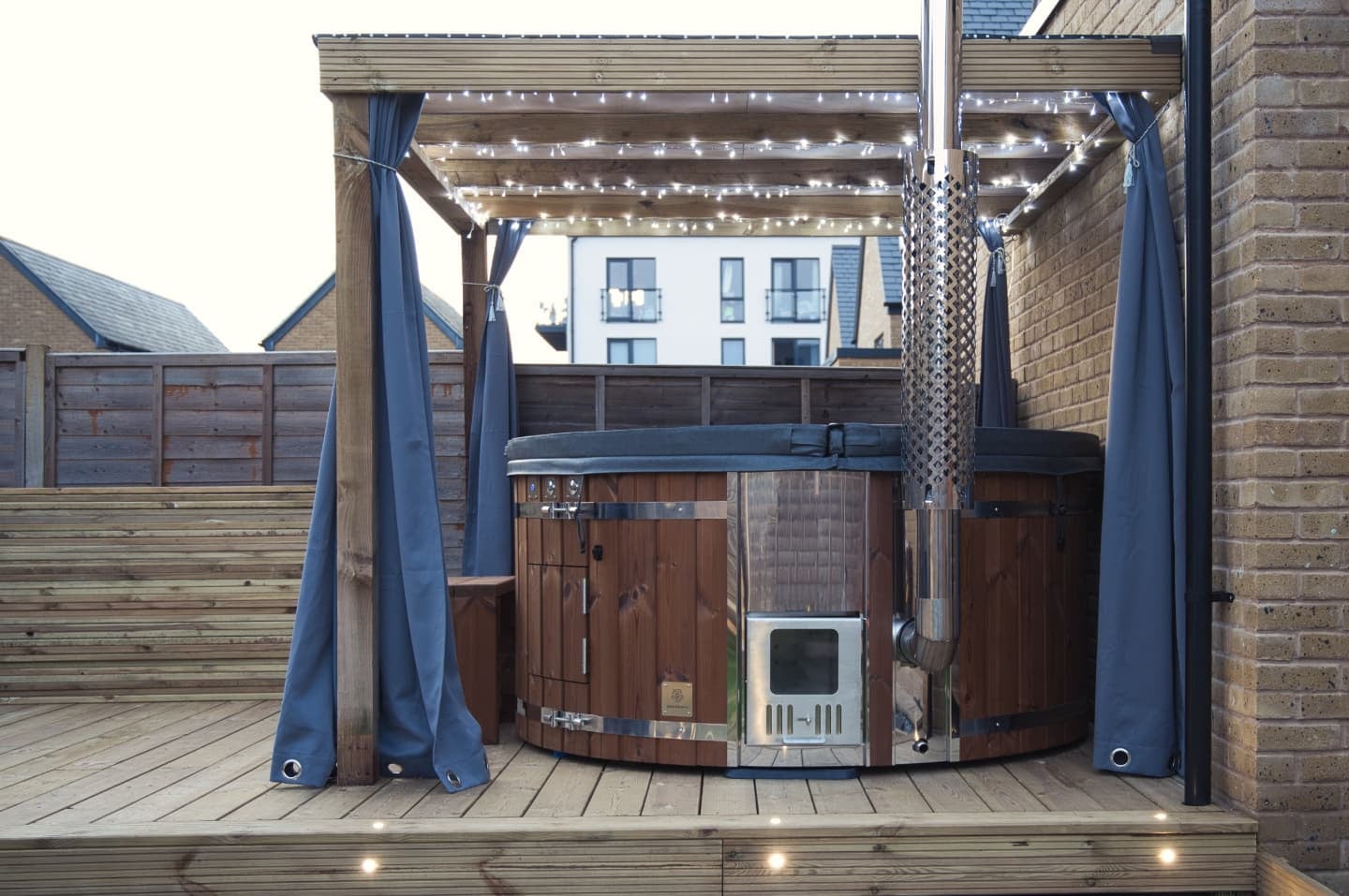 Gardenvity wood fired hot tub on wooden decking under pergola with string lights in a modern courtyard