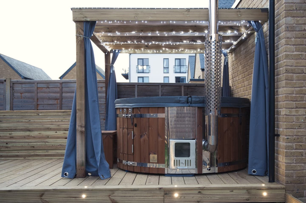 Gardenvity Holzofen-Whirlpool auf Holzterrasse unter Pergola mit Lichterketten in einem modernen Innenhof.