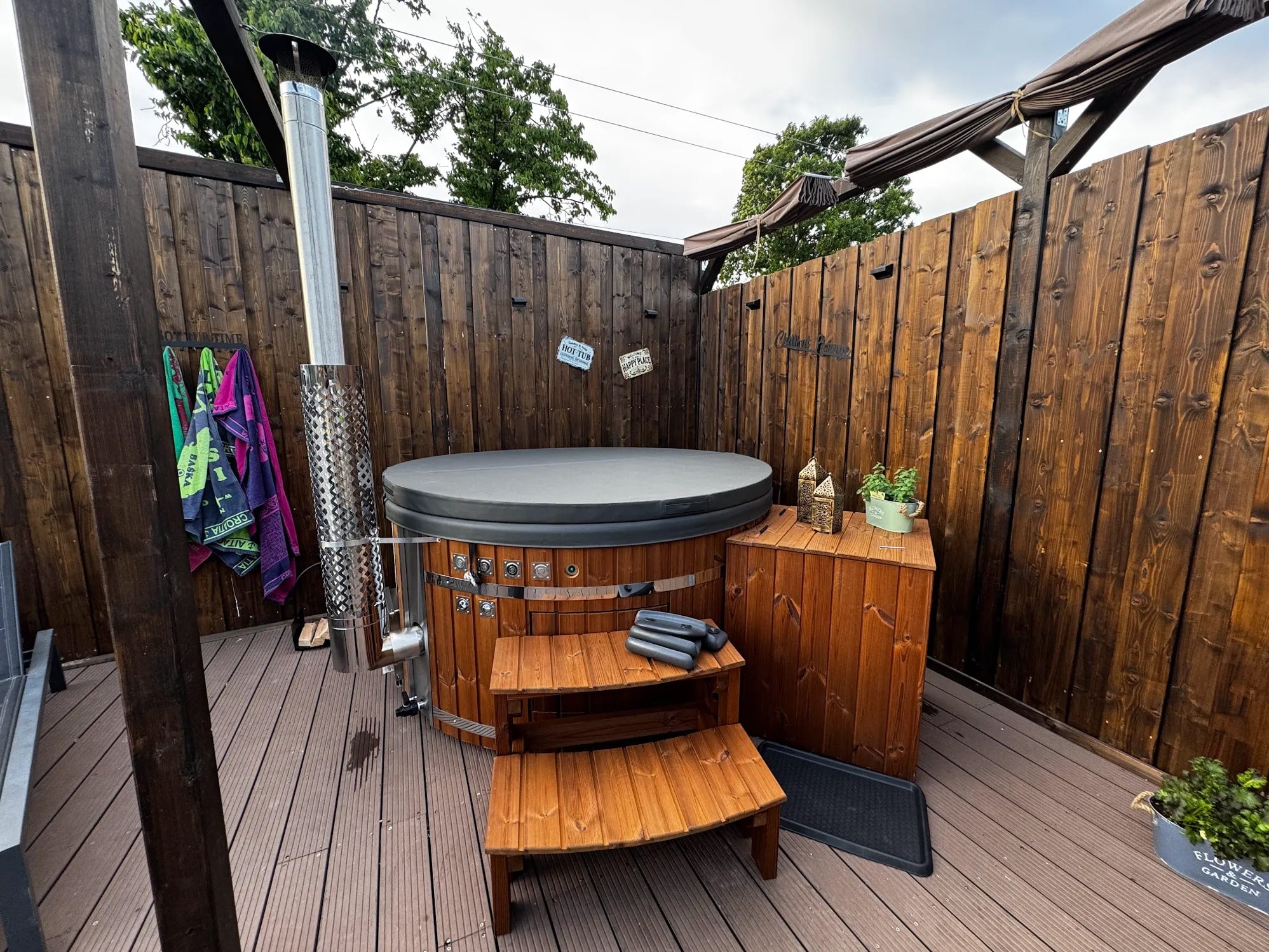 Gardenvity wood fired hot tub on compact patio with dark wood privacy wall and stairs.