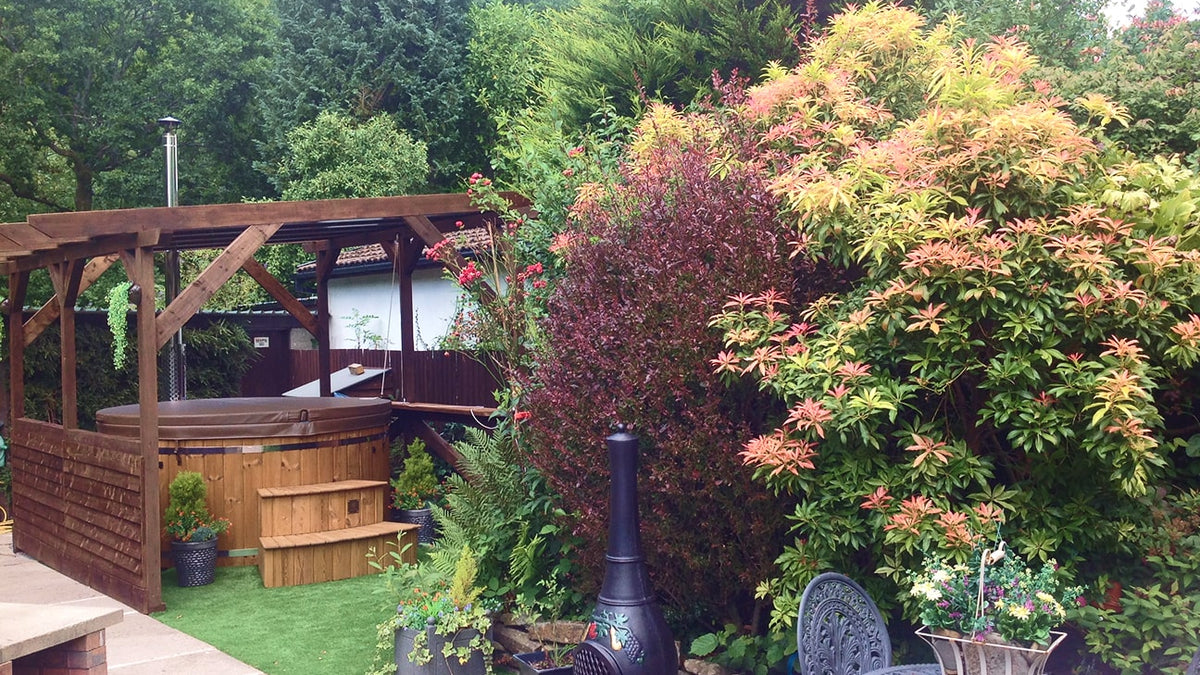 Gartenterrasse mit Holzpergola, Whirlpool, üppigen Sträuchern, Außenkamin und Metalltisch mit Stühlen.