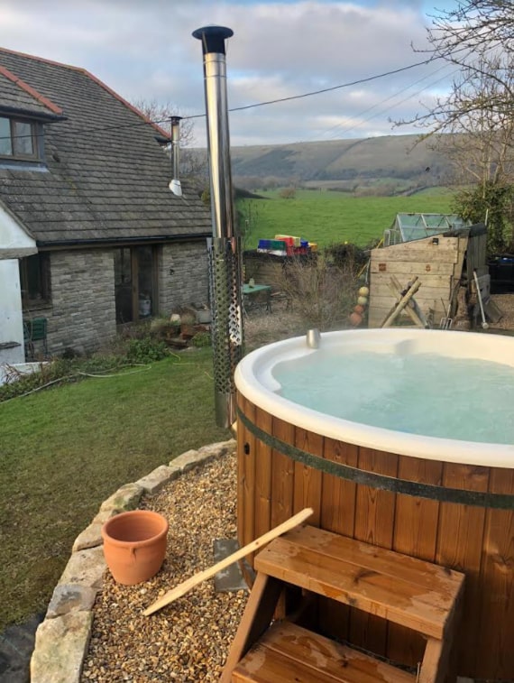 Außenansicht eines runden, holzbefeuerten Hot Tubx mit integriertem Edelstahlofen und Holzstufen neben einem Steinhaus