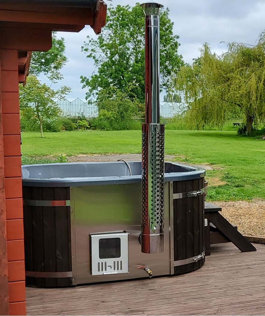 Außenansicht eines hölzernen Hot Tubx mit Metallofen, Edelstahlkamin und Holzstufen auf einer Holzterrasse