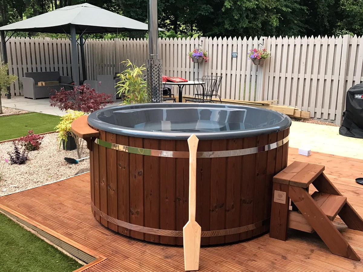 Außenansicht eines runden hölzernen Whirlpools mit Metallbändern, Kamin, Holzstufen und Paddel auf einer Holzterrasse