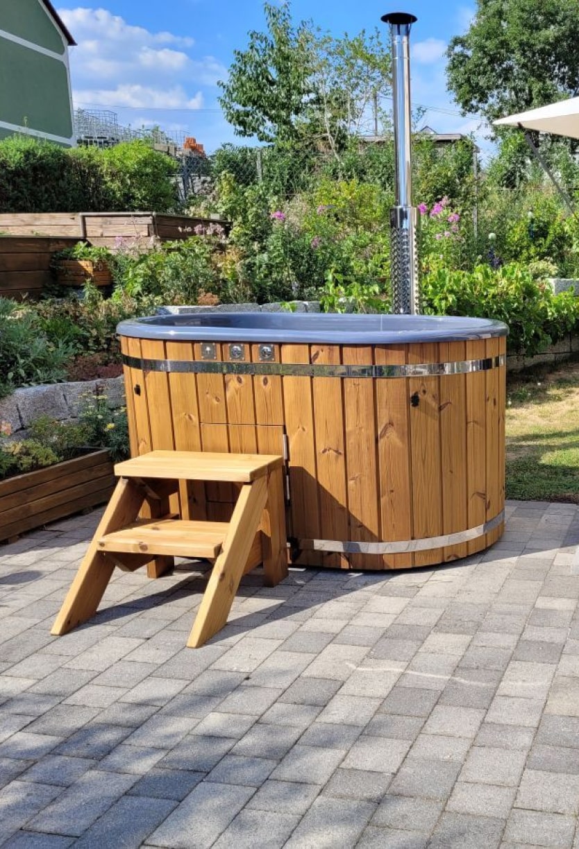 Außenansicht eines hölzernen Whirlpools mit Metallbändern, Schornsteinrohr und Holzstufen auf einer gepflasterten Terrasse im Garten.