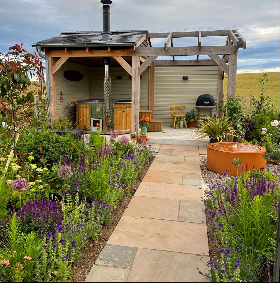 Blick in den Garten mit Holzpergola, Whirlpool, Steinweg und blühenden Pflanzen, die den Bereich umgeben