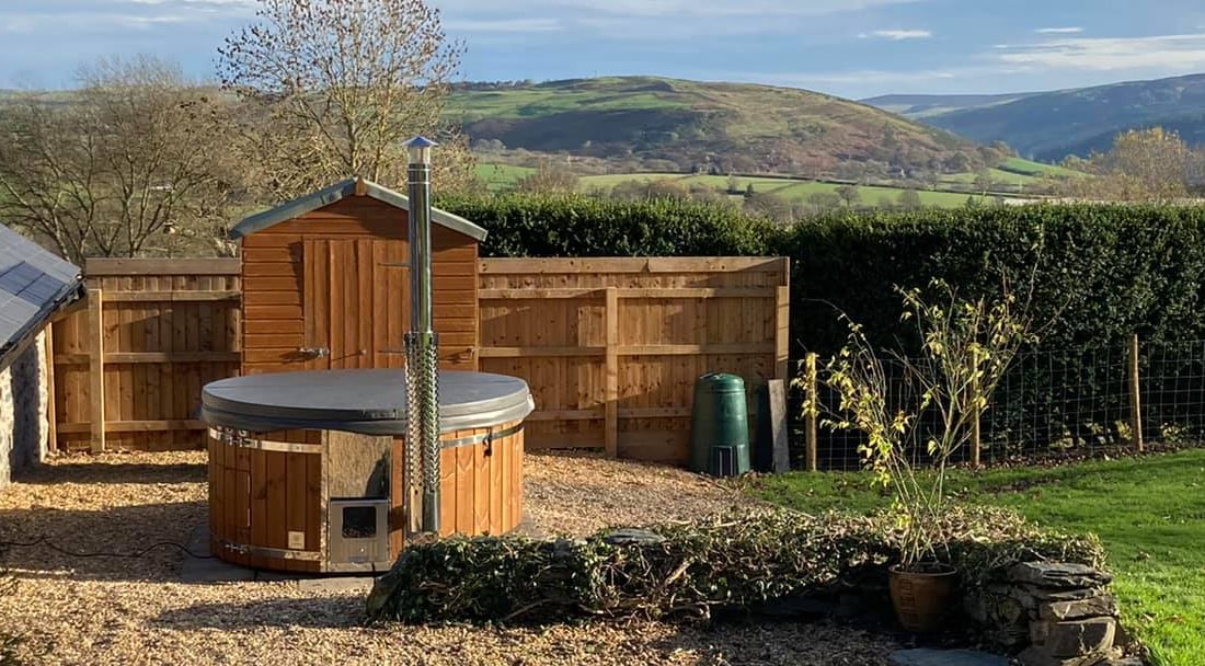 Gardenvity wood fired hot tub installed in garden with wooden fence and scenic hill backdrop