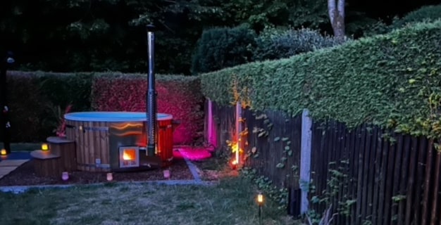 Ein holzbefeuerter Außen-Whirlpool mit eingeschaltetem Ofen und farbenfroher Gartenbeleuchtung, umgeben von Grün und einem Holzzaun in der Abenddämmerung