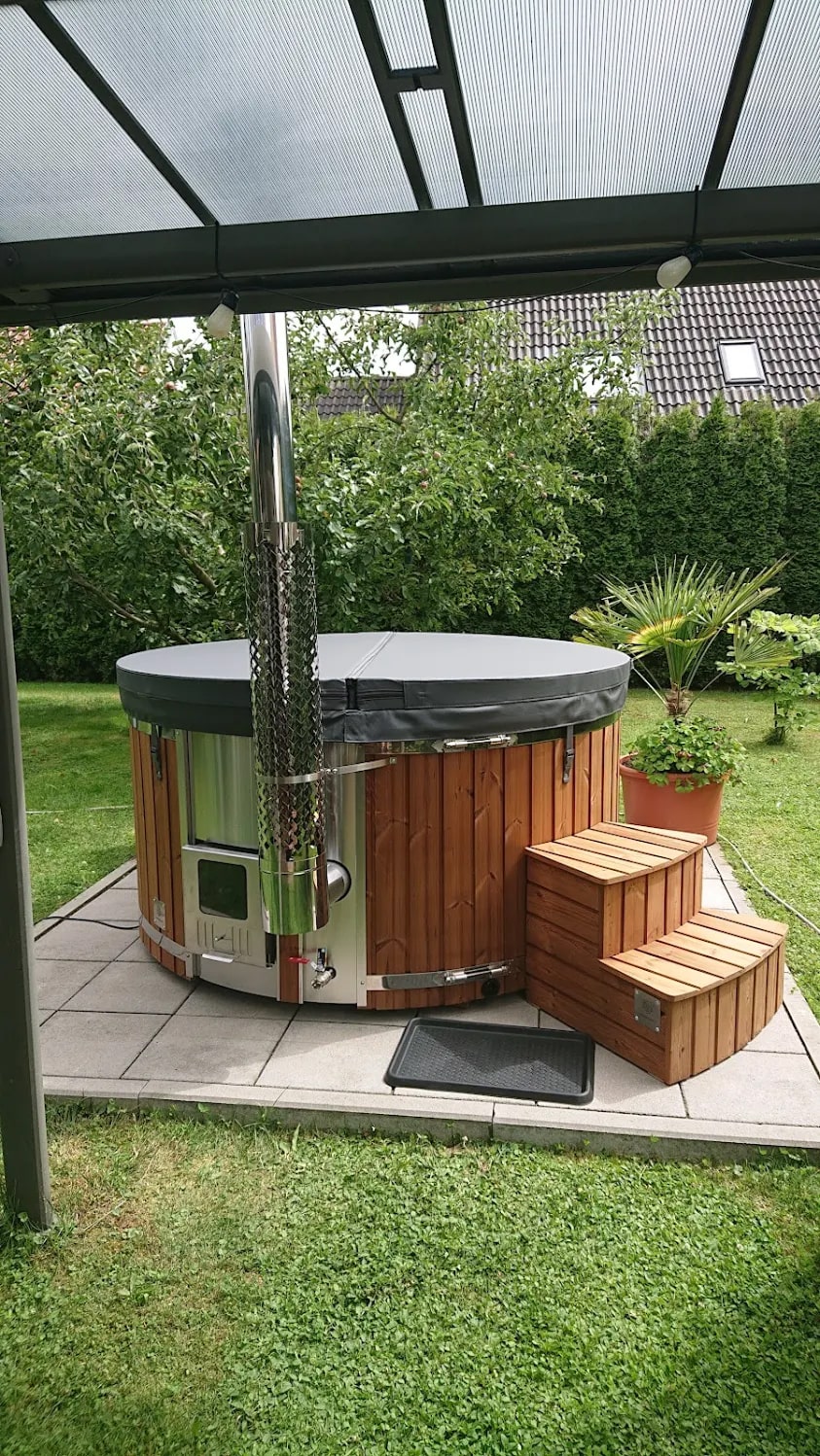 Der kompakte, holzbefeuerte hot tub Gardenvity steht unter einem Terrassendach in einem grünen Garten.