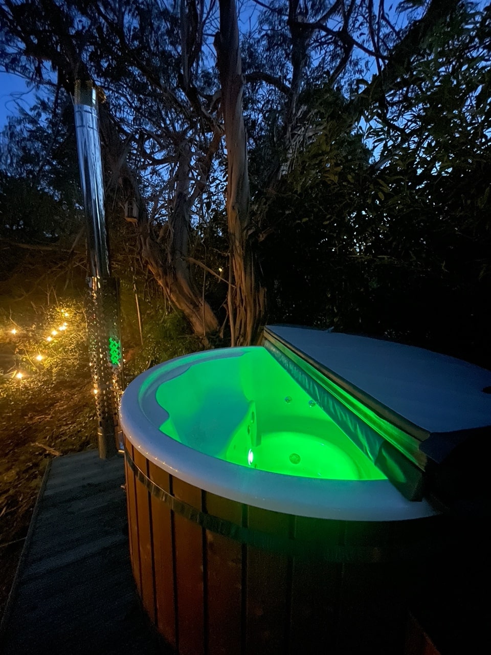 Außenansicht eines holzbefeuerten Whirlpools, der mit grünen LED-Leuchten beleuchtet und von Bäumen und Lichterketten umgeben ist, in der Abenddämmerung.